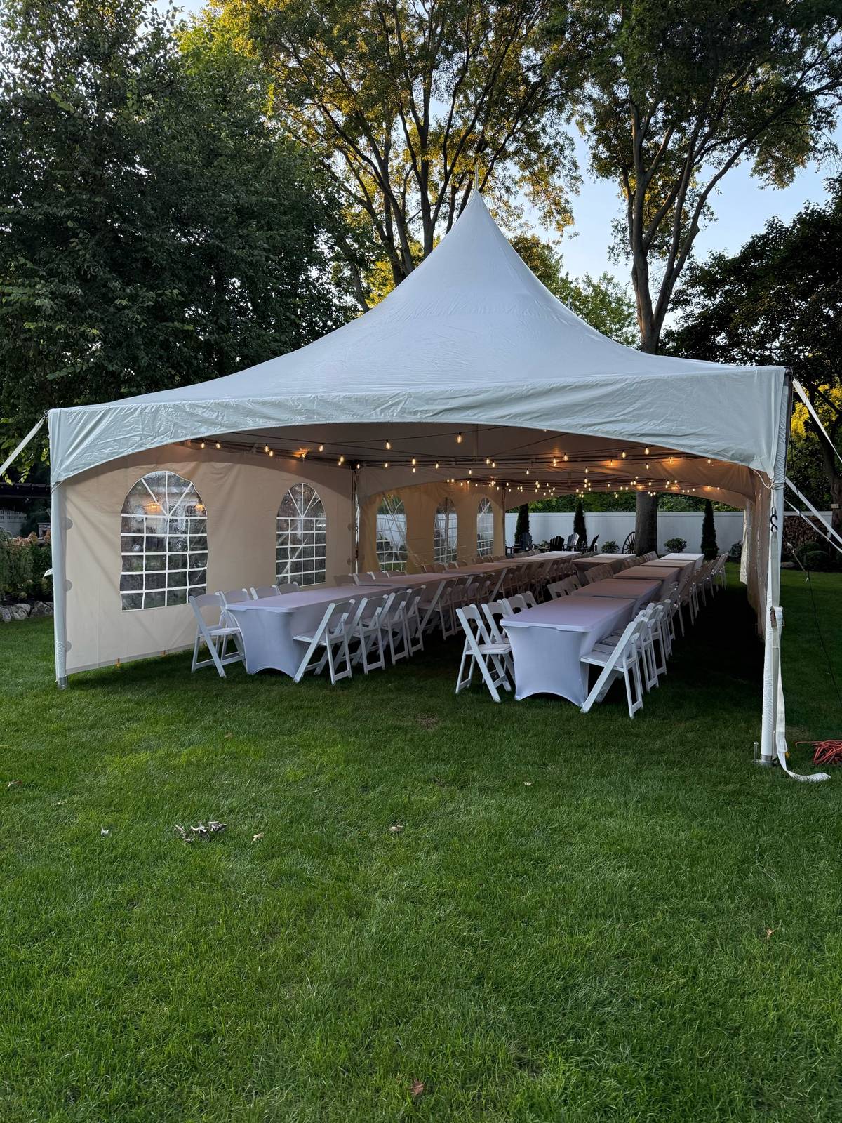View into a high peak tent with long tables, white folding chairs and string lights